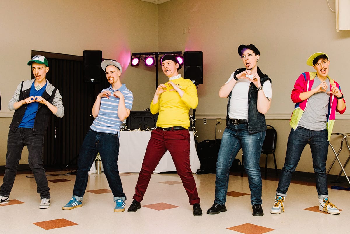 Kelly Prizel photo of five people dressed in jeans and baseball caps in a row with their hands making a heart shape in front of their chests while performing a dance routine at a wedding reception.