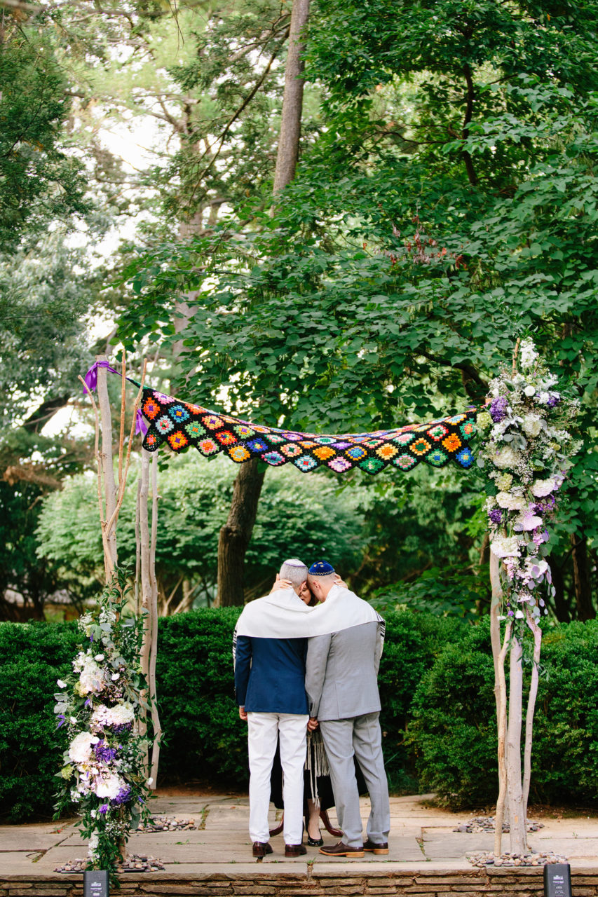 Kelly Prizel photo of two grooms standing under a crocheted chuppah with trees in the background.