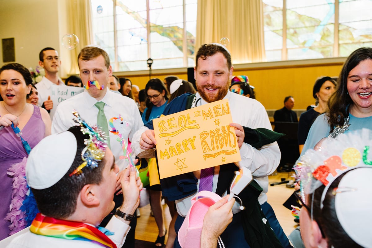 Kelly Prizel photo of a bearded man wearing a yarmulke holding up a sign that says "Real men marry rabbis" in the middle of a crowded dance floor at a wedding receiption