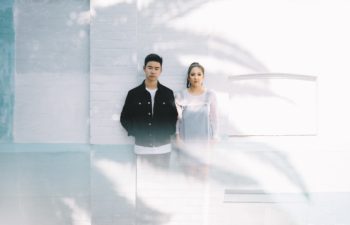 A man and a woman standing against a white wall in the shadow of a tree. Both look at you with serious expressions.