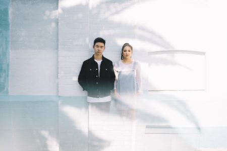 A man and a woman standing against a white wall in the shadow of a tree. Both look at you with serious expressions.