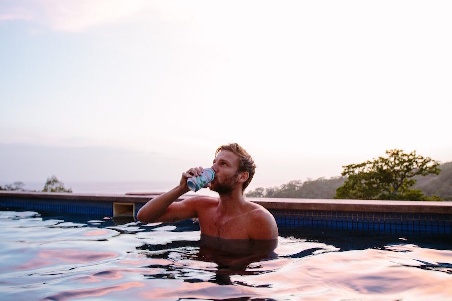 A man drinks a beer while sitting in a pool at a pink and blue sunset