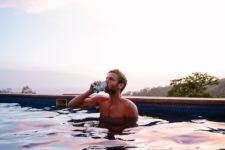A man drinks a beer while sitting in a pool at a pink and blue sunset