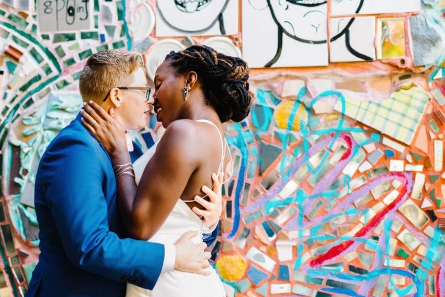 A mixed-race wedding couple kisses in front of a colorful wall