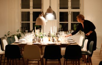 A blond woman in a black dress sets a large table for a feast under pendant lights in front of tall windows