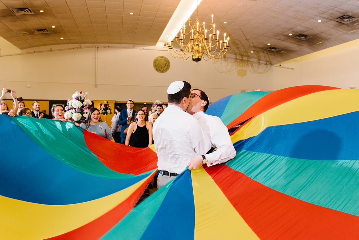 Kelly Prizel photo of two grooms kissing on the dance floor of a wedding reception with a brightly colored parachute swirls around them and guests look on
