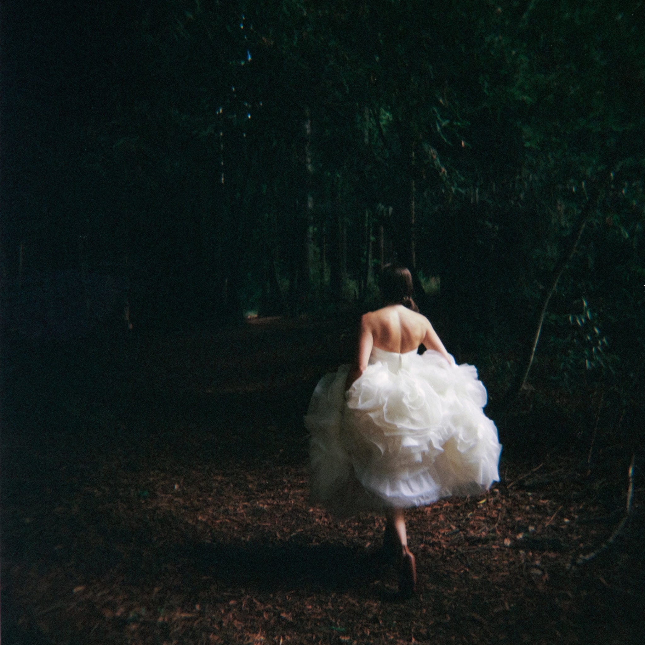 A woman gathers her full, ruffled wedding dress skirt around her and walks into the dark woods