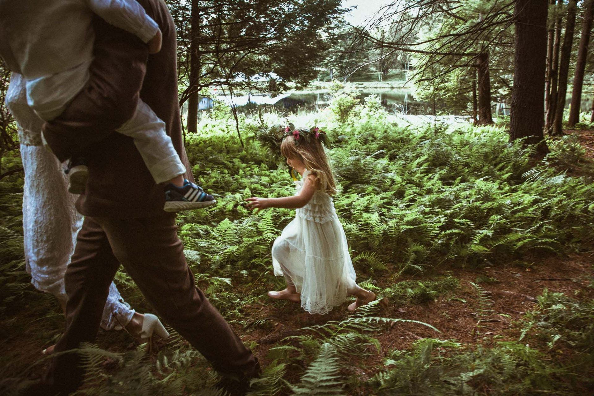 a small girl in a white dress and flower crown walks through the green woods behind a bride and a groom carrying another small child