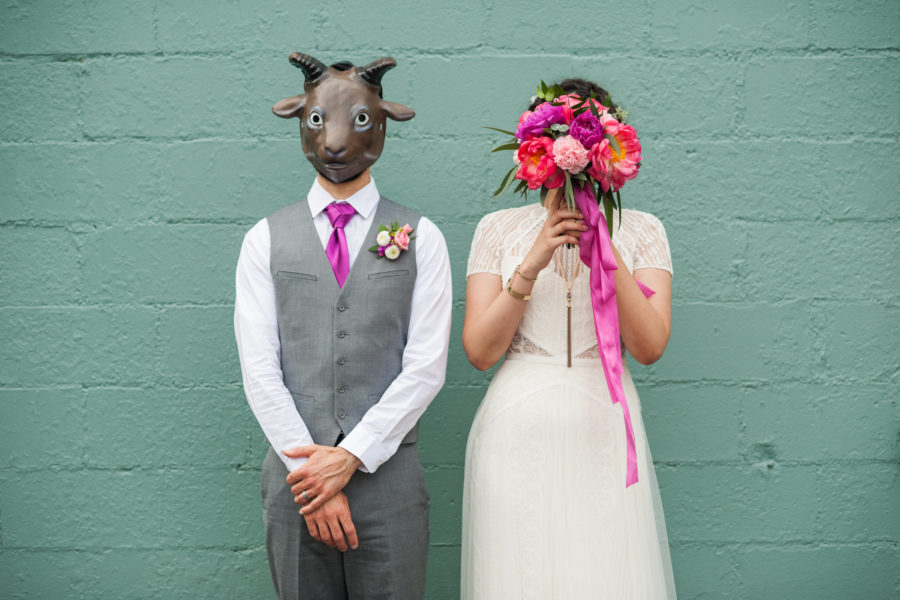 A bride and groom in wedding clothes stand in front of a teal wall. The groom wears a goat mask and the bride holds her brightly colored bouquet in front of her face.