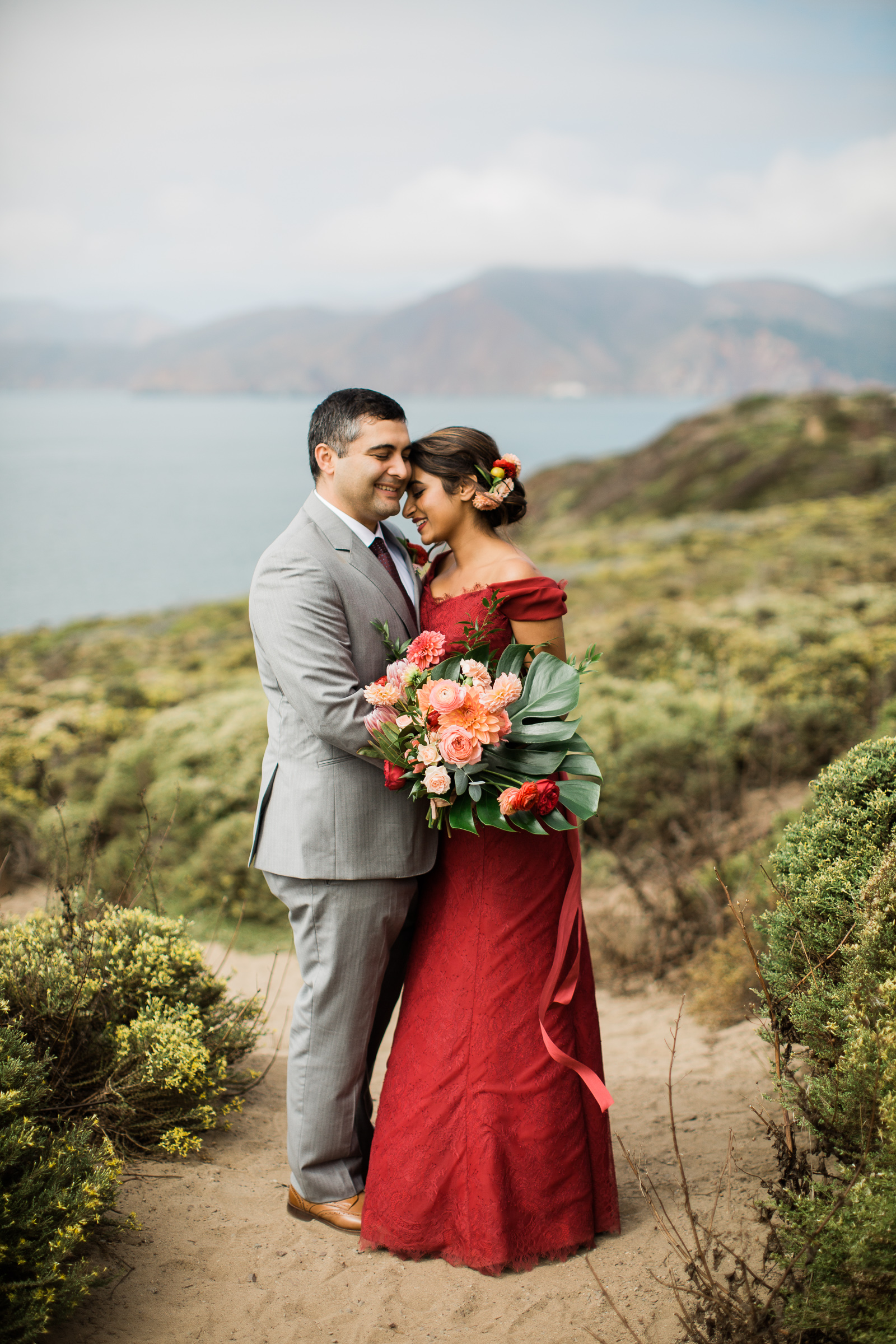 A man in a grey suit and woman in a red wedding gown embrace while standing on a hilltop overlooking the ocean in a Manali Anne photo