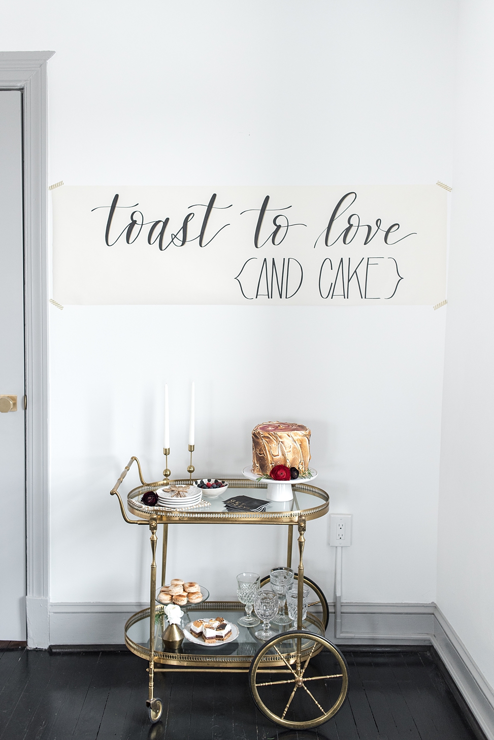 A sign reads "toast to love {and cake}" above a bar cart set with a bruleed cake, taper candles, and other treats at Bureau co-working and event space