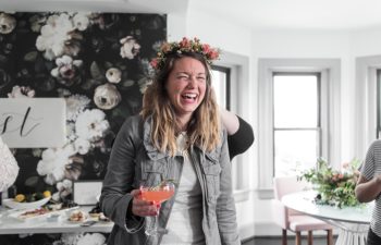 A woman holding a large pink cocktail laughs while her flower crown is adjusted by someone standing behind her in BUREAU's lounge area