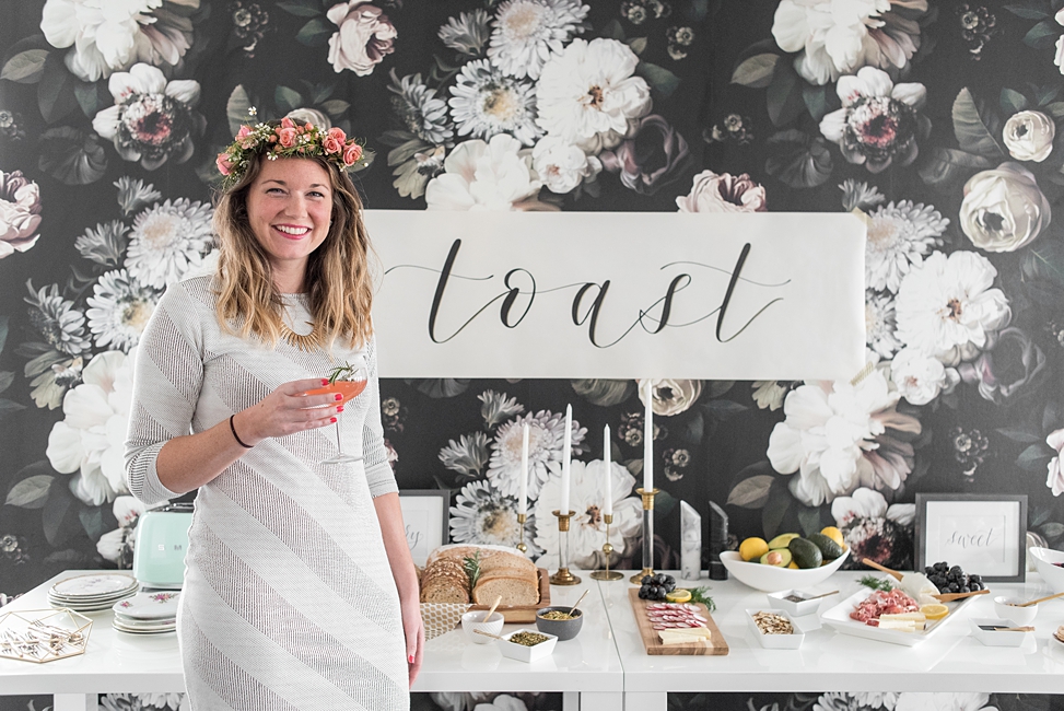 A woman wearing a flower crown and holding a champagne glass smiles as she stands in front of a sign that reads "toast" which hangs above a tasty looking build your own toast bar at Bureau co-working and event space.