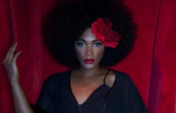A woman in a black lingerie set with a silky black robe sits in a red velvet chair and looks at you. She has two red flowers in her natural hair