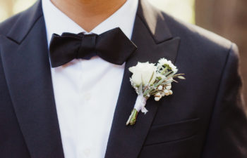 The torso of a man in a black tux with bow tie, wearing a white floral boutonnière.