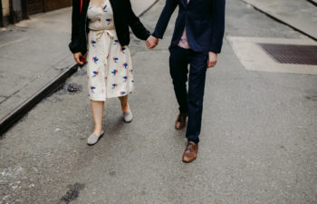 A woman in a white floral print dress with a black sweater holds hand with a man in a black suit on a grey street