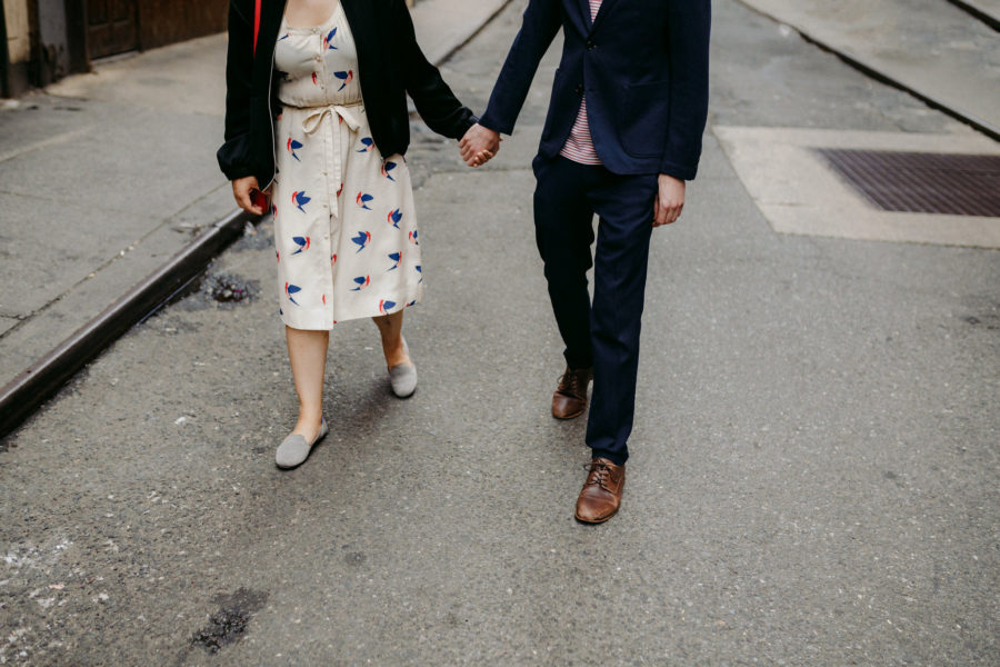 A woman in a white floral print dress with a black sweater holds hand with a man in a black suit on a grey street