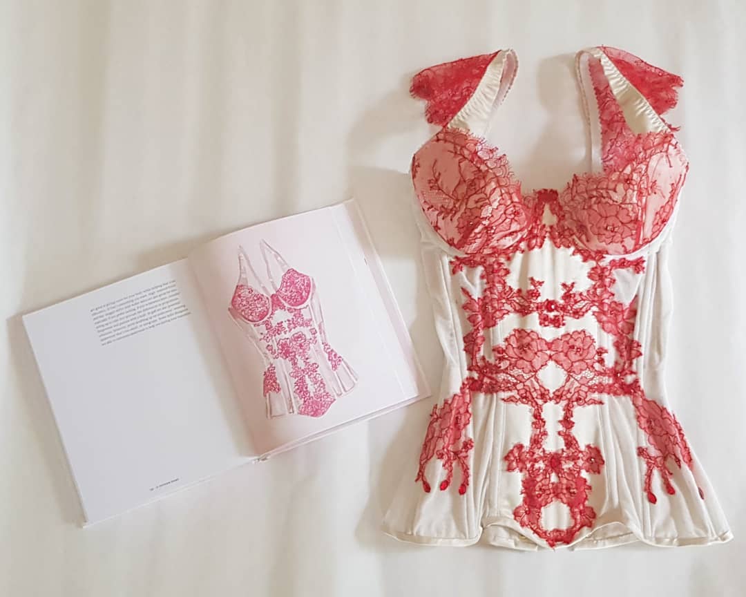 The book In Intimate Detail is open to a page featuring a watercolor illustration of a red embroidered white corset. The book sits next to the original inspiration for the illustration on a white fabric background, an example of honeymoon lingerie.