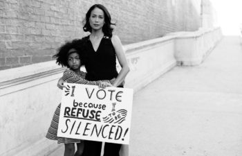 A woman and a young girl embrace and hold a sign that reads, "I vote because I refuse to be silenced!"
