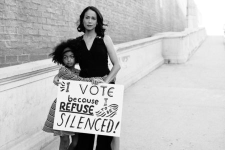 A woman and a young girl embrace and hold a sign that reads, "I vote because I refuse to be silenced!"