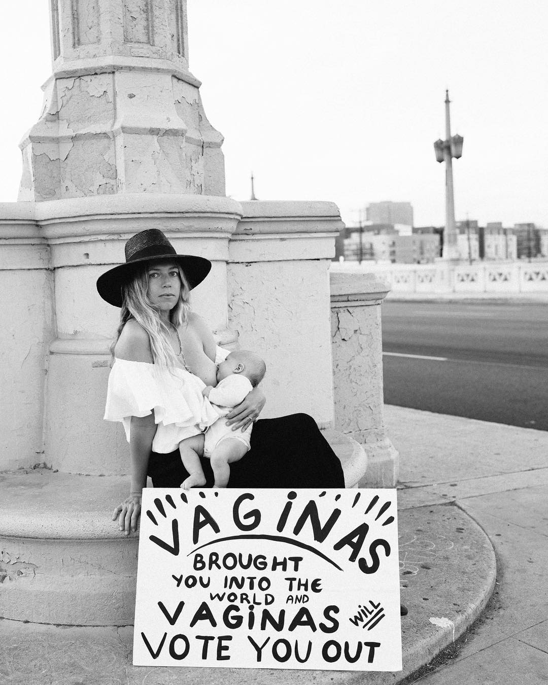 A woman in a wide-brim hat looks at you while sitting at the base of a column, breastfeeding her baby. A sign leans against her that reads, "Vaginas brought you into this world and vaginas will vote you out."
