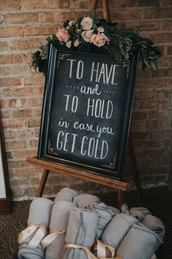 Winter wedding ideas for cozy hospitality for guests—Chalkboard sign in front of a brick wall that reads "To have and to hold in case you get cold" above a basket of grey fleece blankets