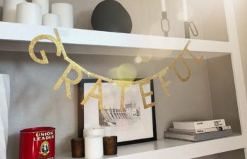 A gold glittery banner reading "Grateful" hangs from a white shelf with candles and books and pictures.
