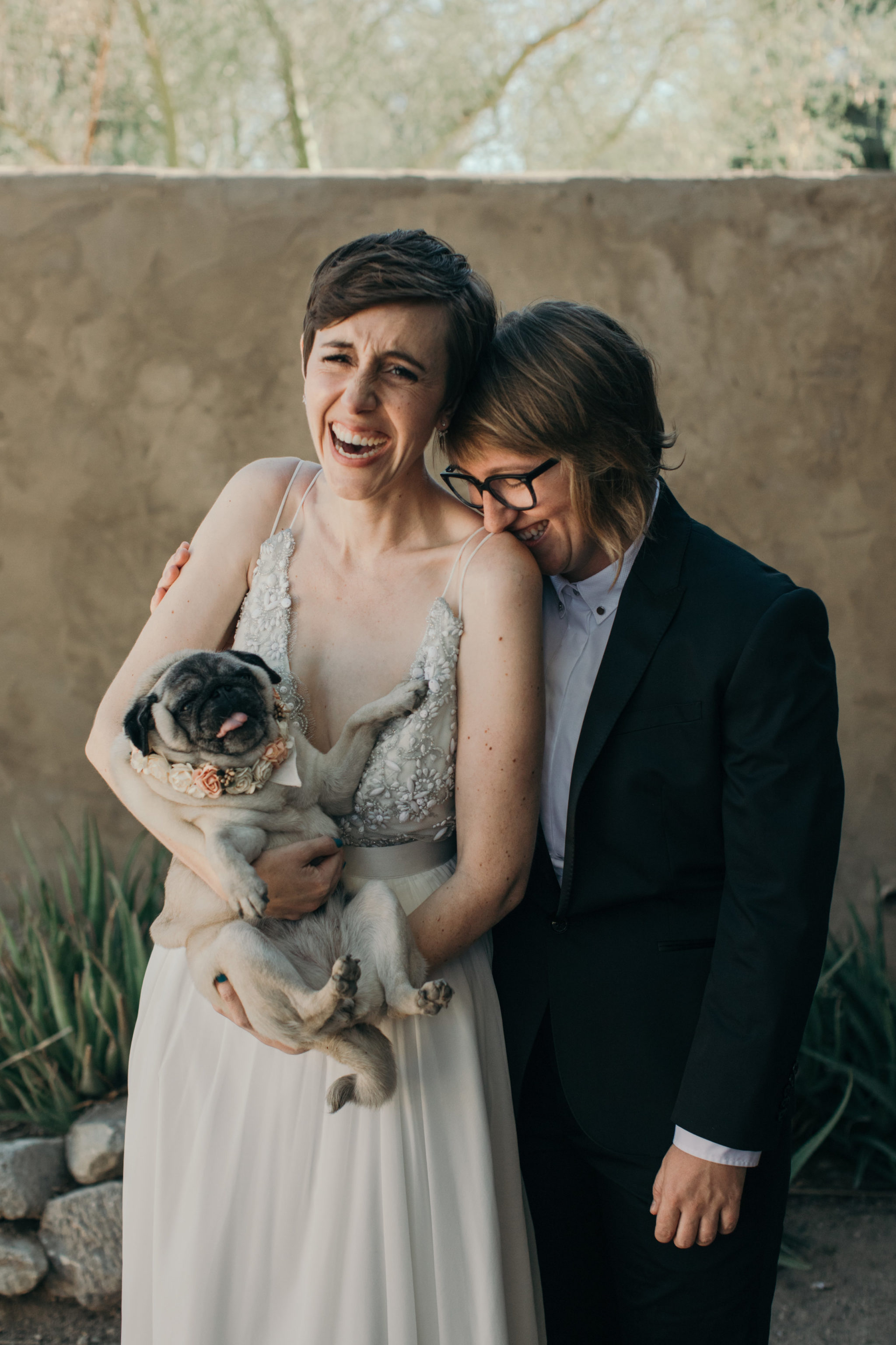 A queer wedding couple is embracing and laughing as one holds their pug who's wearing a floral garland and squirming to be put down in a photo by Betty Clicker