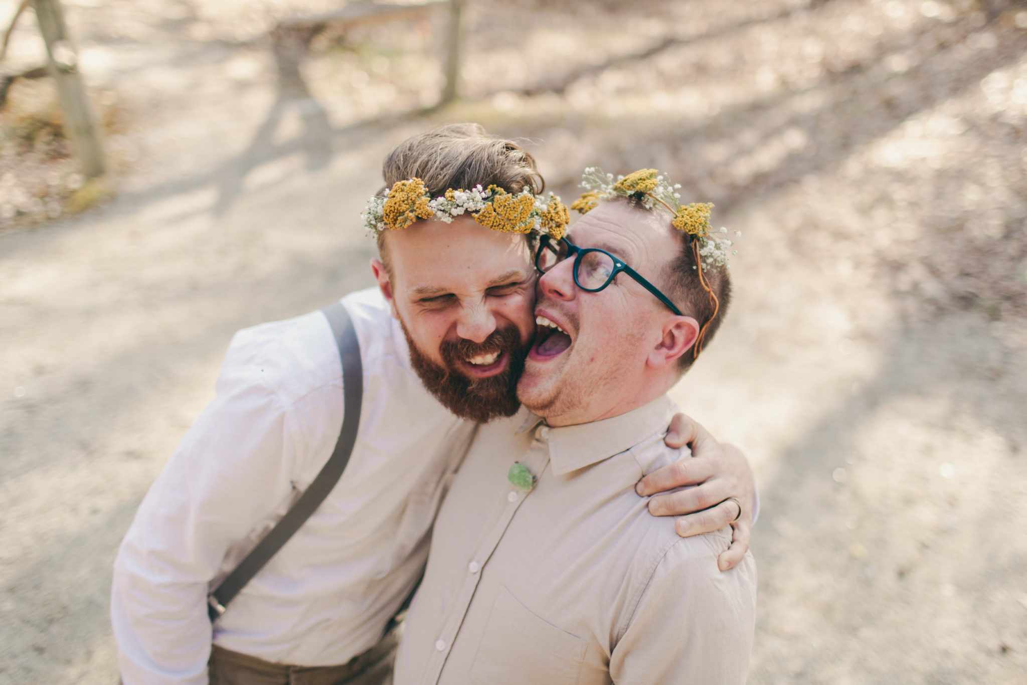 Two grooms in tan shirts and yellow flower crowns laugh and embrace in a photo by Betty Clicker