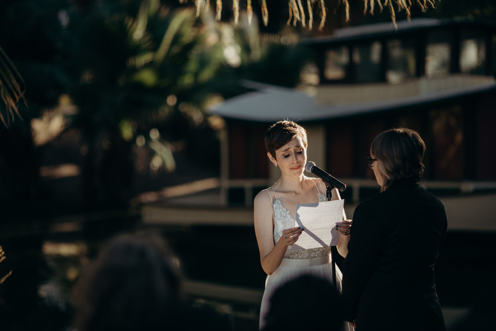 Bride with short hair and beaded gown is tearing up as she faces her partner and reads her vows in a photo by Betty Clicker