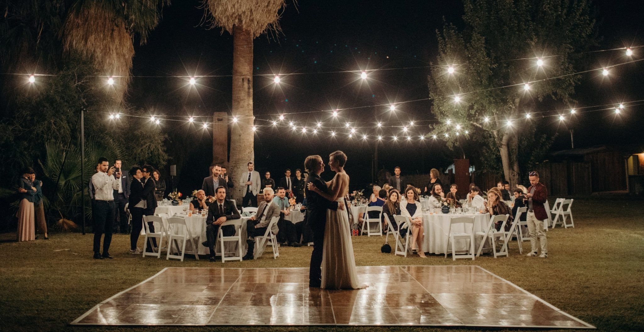 A wedding couple has their first dance on a outdoor dance floor surrounded by palm trees and string lights as their guests look on in a photo by Betty Clicker