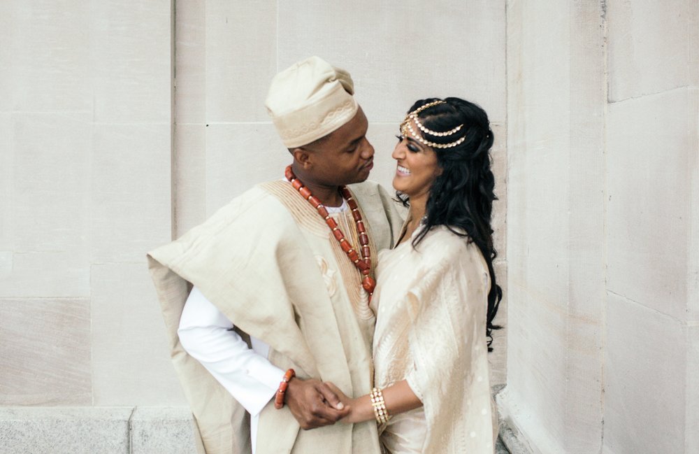 A bride in a traditional Indian wedding outfit and a groom in traditional Nigerian wedding outfit hold hands and smile at each other in a photo by Betty Clicker