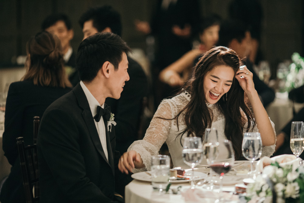 An Asian bride is sitting at a sweetheart table with her groom and she tucks her hair behind her ear as she laughs and reaches a hand out to groom in a photo by Betty Clicker