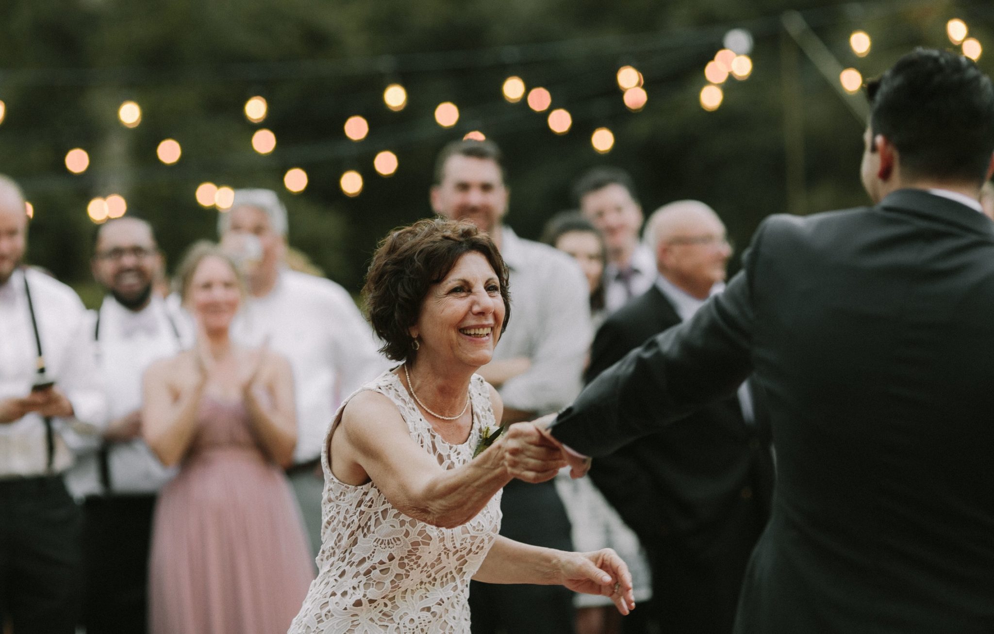 A smiling older woman in a white lace dress reaches out to a man on a dance floor lit by edison bulbs as guests look on in a photo by Betty Clicker