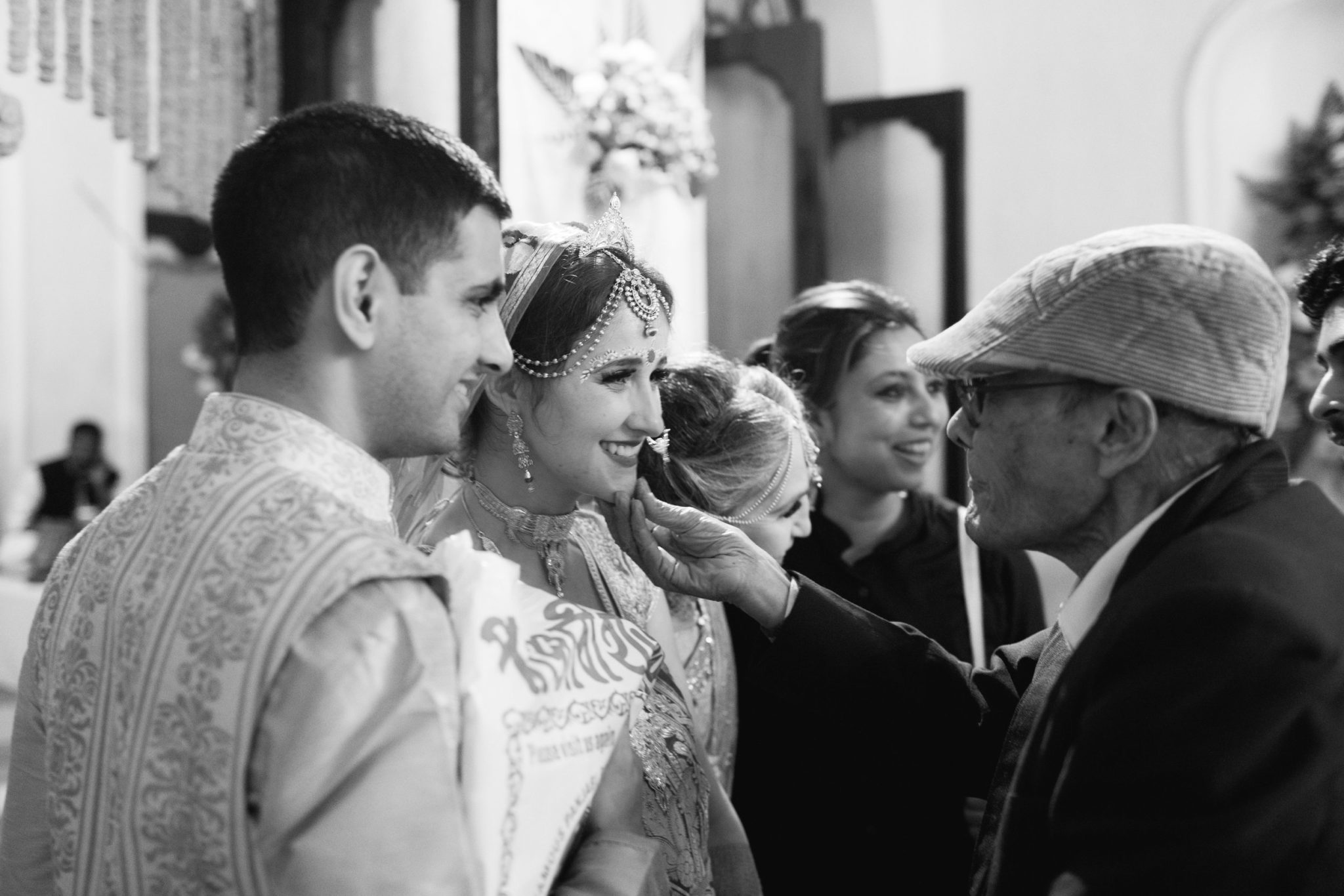 An elderly man greets the bride and groom, who are dressed in traditional Indian wedding clothes, and reaches out to touch the bride's face in a photo by Betty Clicker