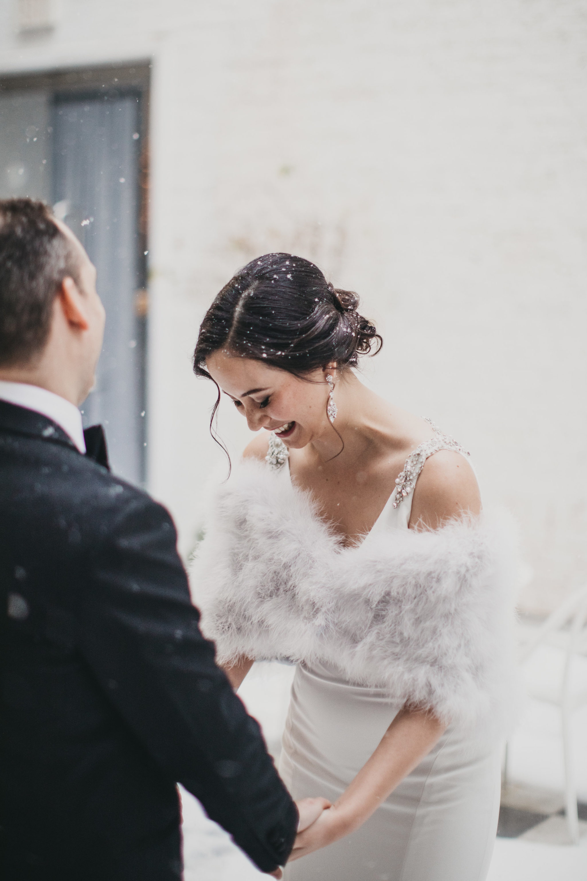 Bride with dark hair and white fur bolero is covered in snow flurries as she looks down and laughs while holding hands with her groom in a photo by Betty Clicker