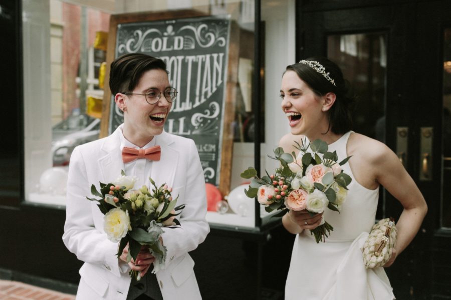 Queer wedding couple in white jacket and pink bow tie and white wedding dress both hold bouquets and laugh excitedly in a photo by Betty Clicker