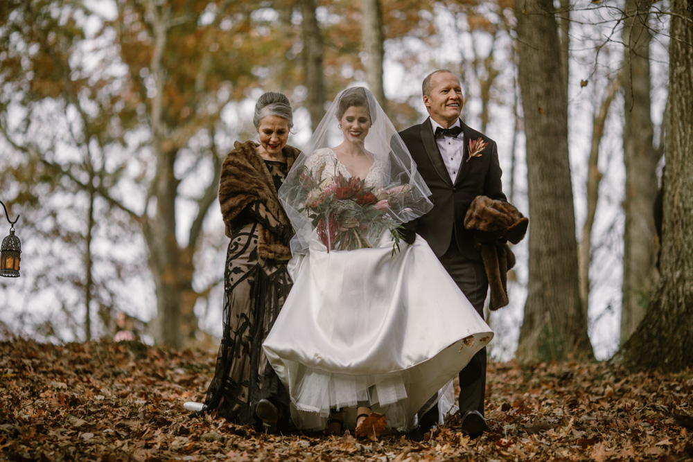 A bride wearing a veil is escorted down a hill covered in fall leaves with her mother in a brown gown and fur on her left, and her father in a suit on her right in a photo by Betty Clicker