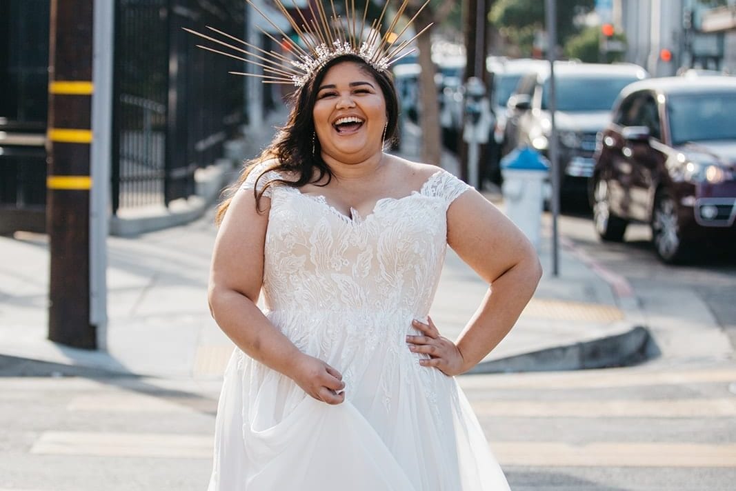 A woman in a radiant crown and plus size wedding dress from Lace and Liberty crosses the street in front of a one way sign, smiling.