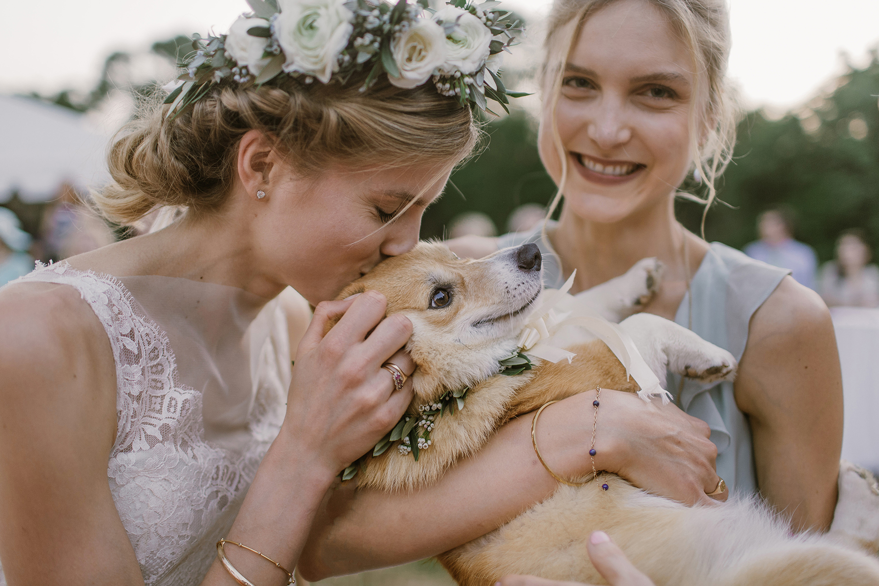 A blond woman in a grey dress looks at you and smiles as she holds a dog, and a woman in a flower crown and a wedding dress kisses the dog's head in a Sarah Gormley photo
