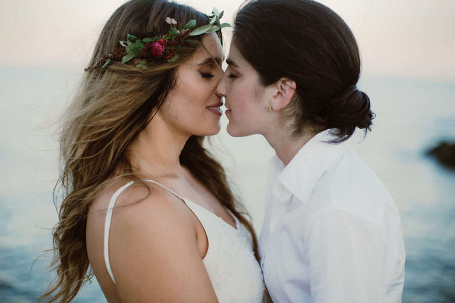Two women in formal white clothes, one wearing a flower crown, kiss in front of the ocean