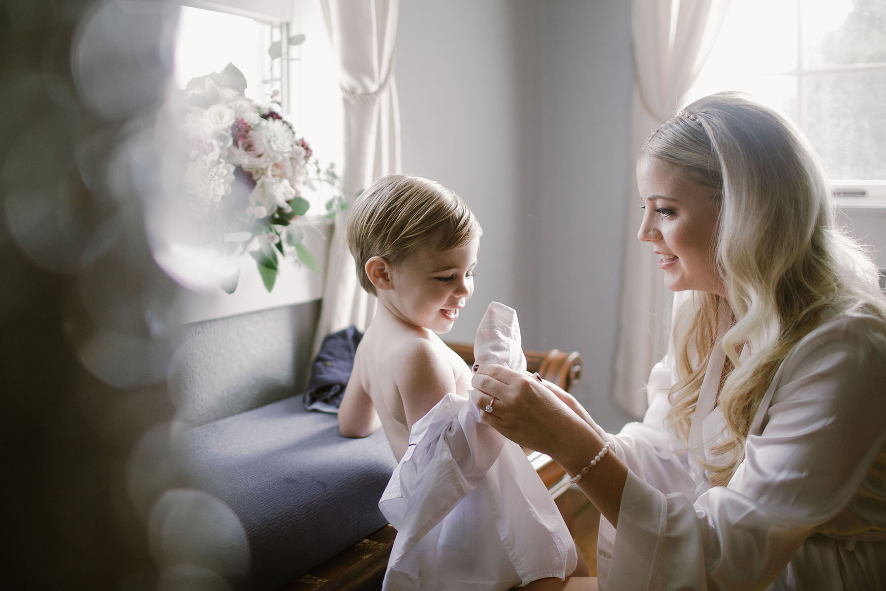 A blond woman helps a small child get a formal shirt on in a brightly lit light room in a Sarah Gormley photo