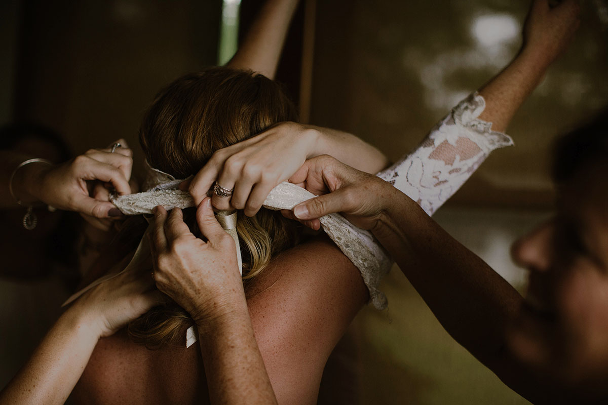 Several sets of women's hands help a bride into her outfit, pulling a lace top over her head in a Sarah Gormley photo