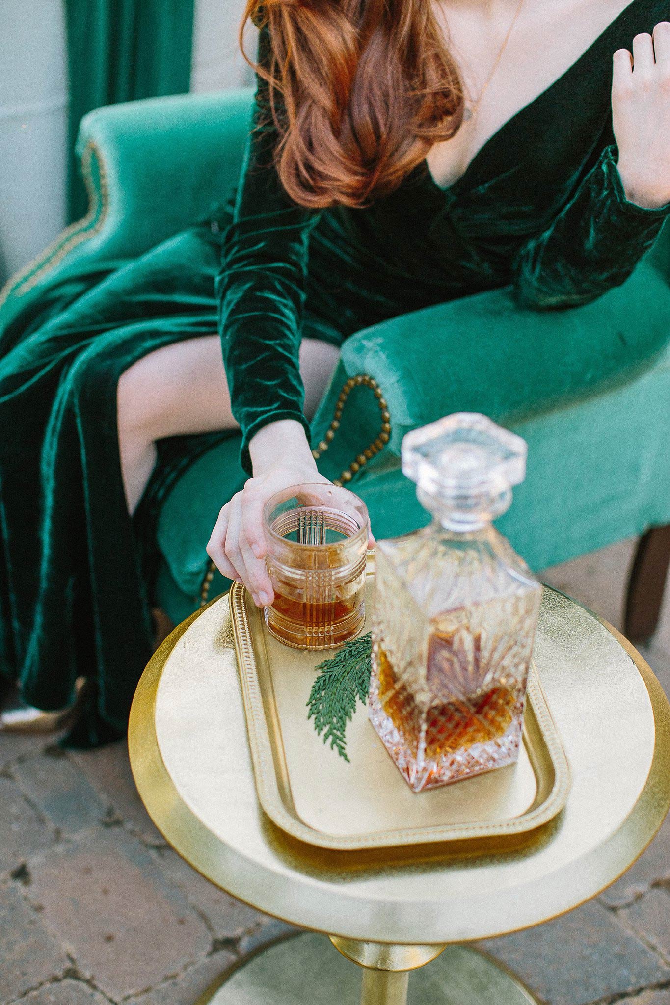 Woman in wearing a winter wedding colors velvet dress in emerald green, sitting in green chair reaching for whiskey glass