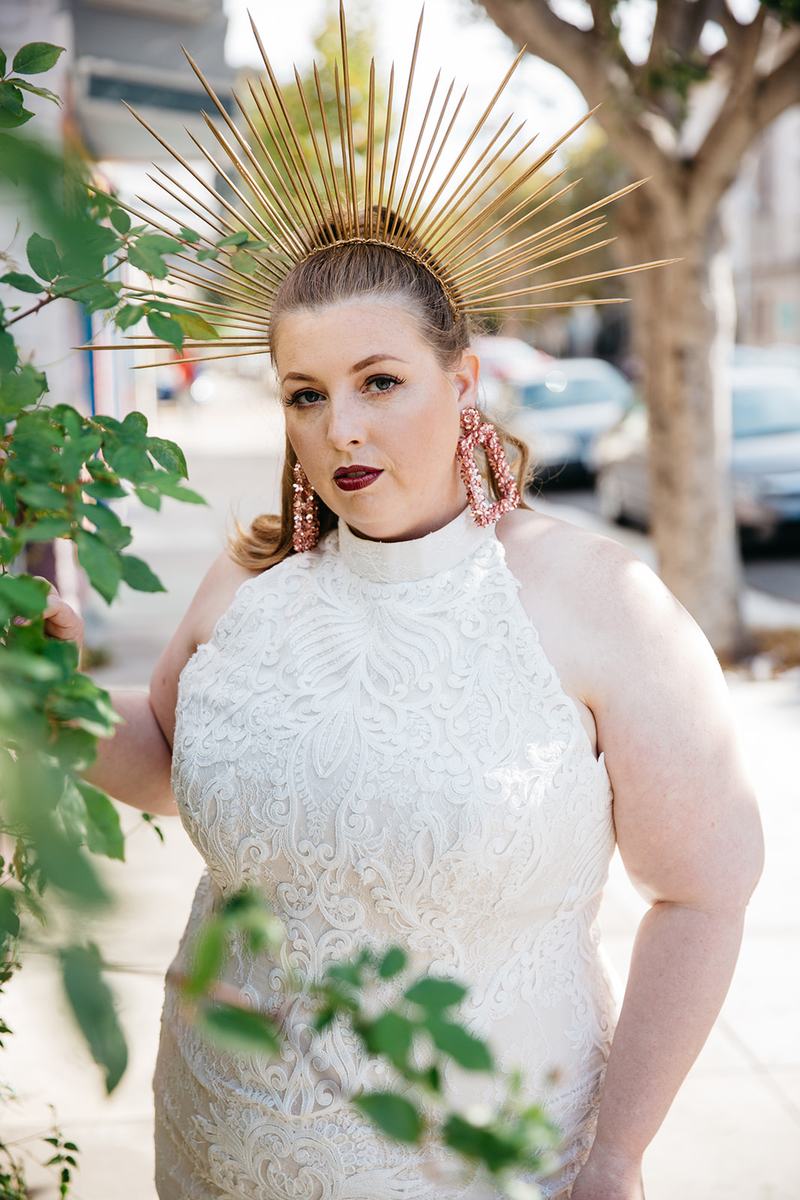 A woman in a Lace and Liberty x APW plus size wedding dress wearing a radiant crown looks at you next to a green plant on the sidewalk