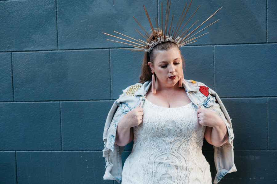 A woman in a radiant gold crown and plus size wedding dress from Lace and Liberty looks down as she pulls over her shoulders a light wash denim jacket covered in patches