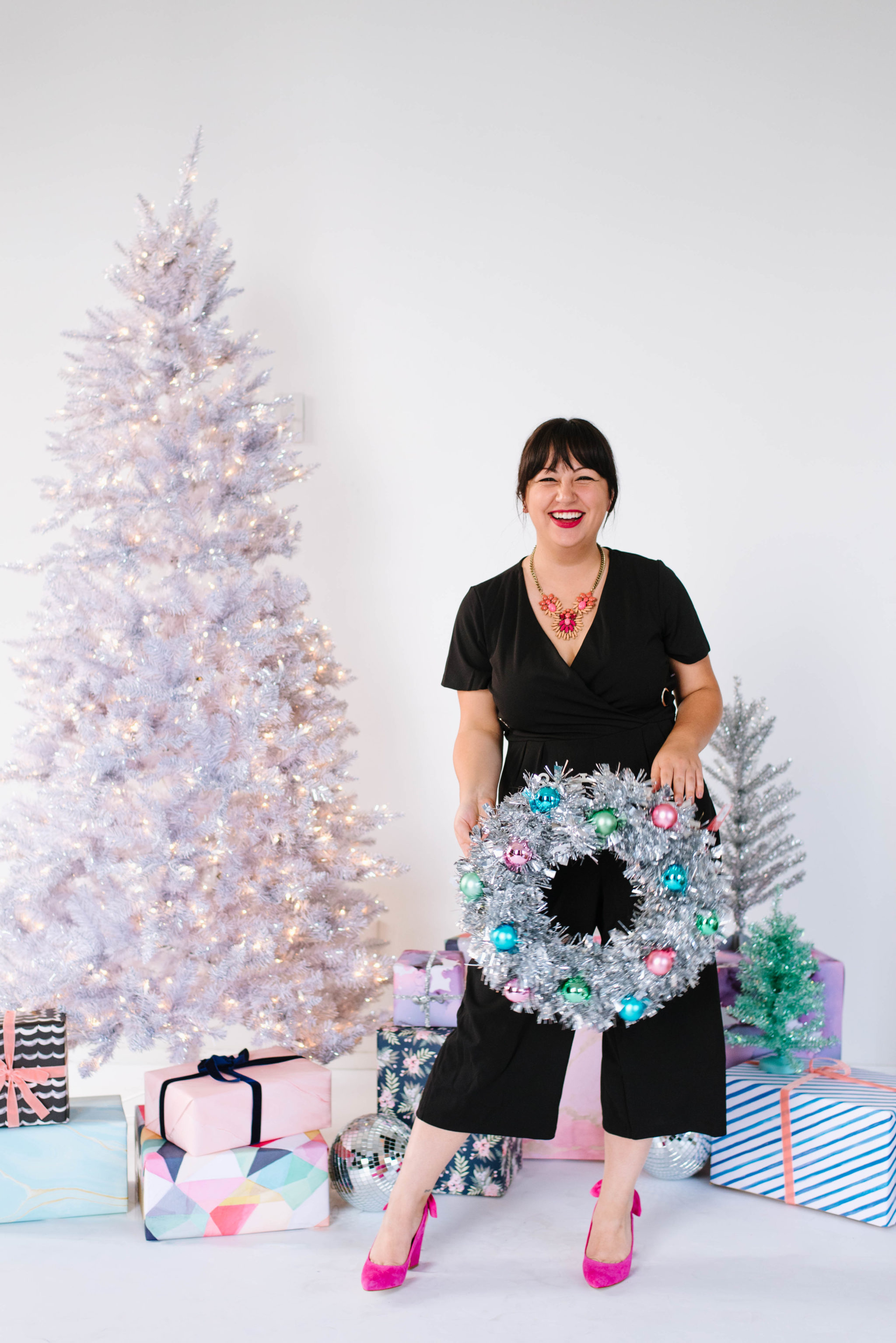 A woman wearing a black jumpsuit from Walmart with a statement necklace and hot pink heels in front of a white Christmas tree and a pile of wrapped Christmas presents