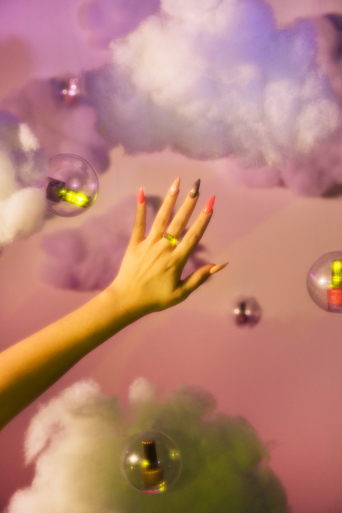 a hand with different nail polish colors on each finger in front of a pink cloud background with floating bubbles of nailpolish
