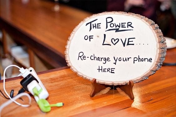 Wedding ideas for guest convenience—a charging station with a power strip and multiple adapters for phones next to a sign written on a slice of wood that reads, "The power of love... re-charge your phone here"