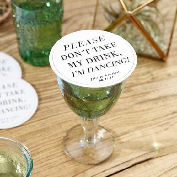 wedding ideas for coasters—Drink Saving Coaster that reads "please don't take my drink, I'm dancing" with the couple's names and wedding date. It sits on a green and clear glass goblet